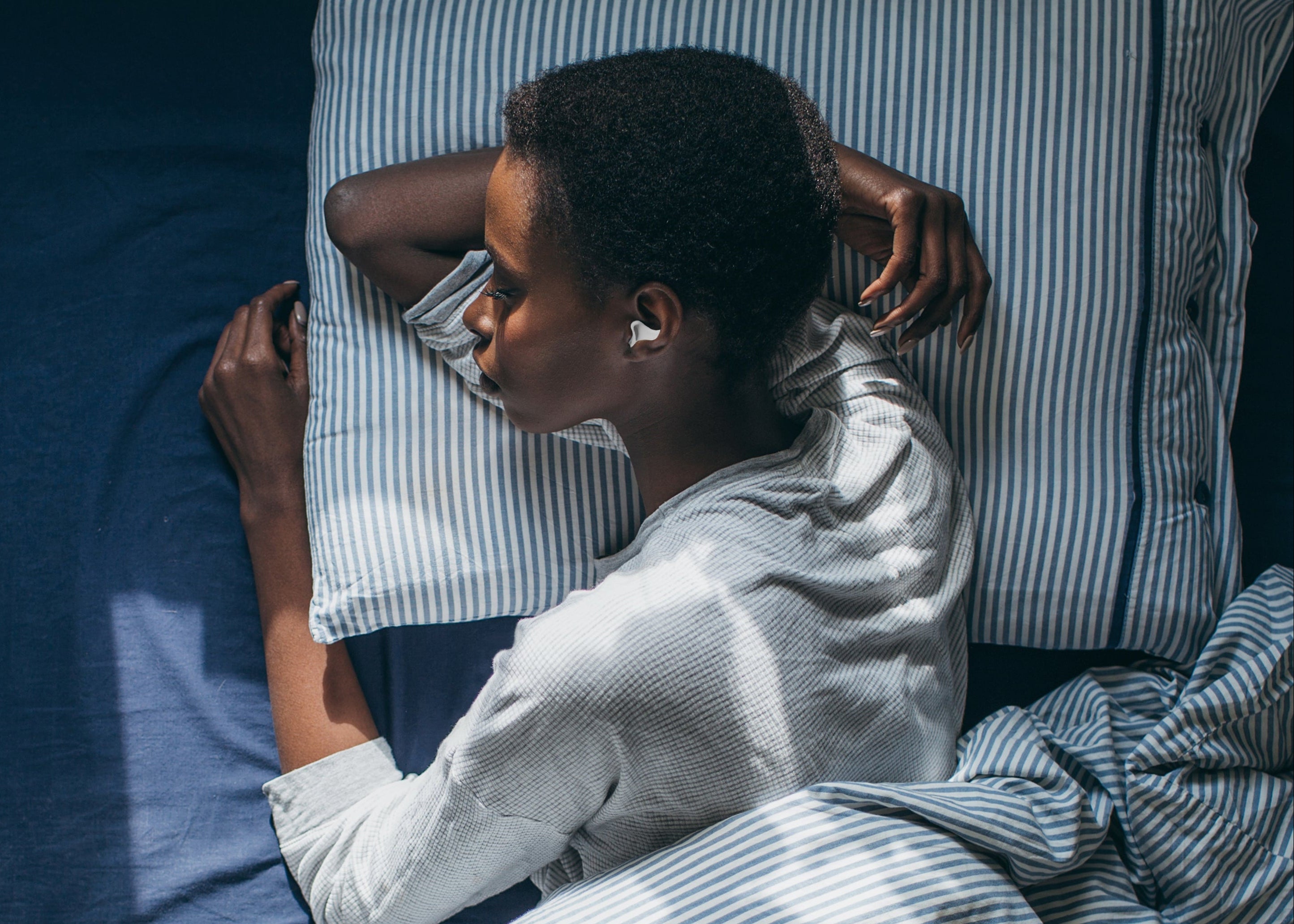 Person liegt schlafend auf einem Bett mit gestreifter Bettwäsche und trägt einen Ohrstöpsel im Ohr.