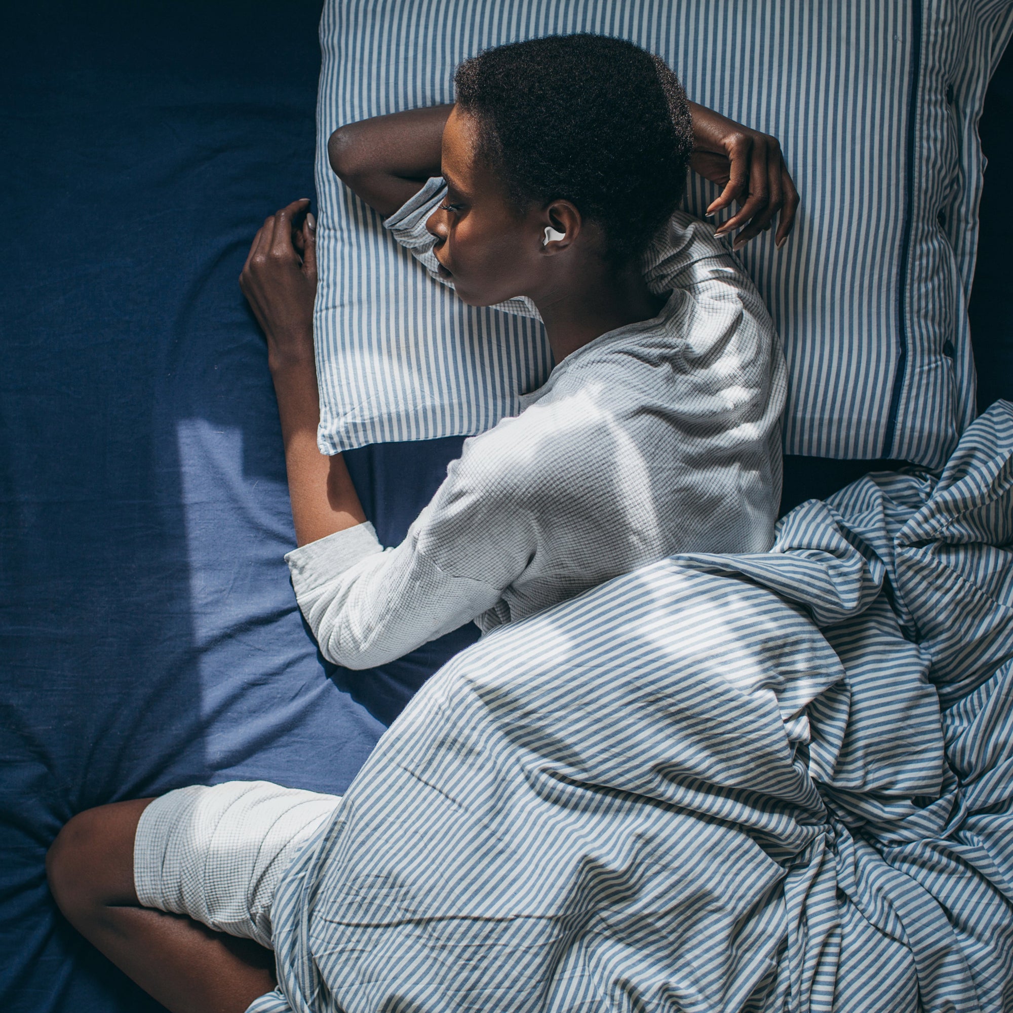 Person liegt schlafend auf einem Bett mit gestreifter Bettwäsche und trägt einen Ohrstöpsel im Ohr.