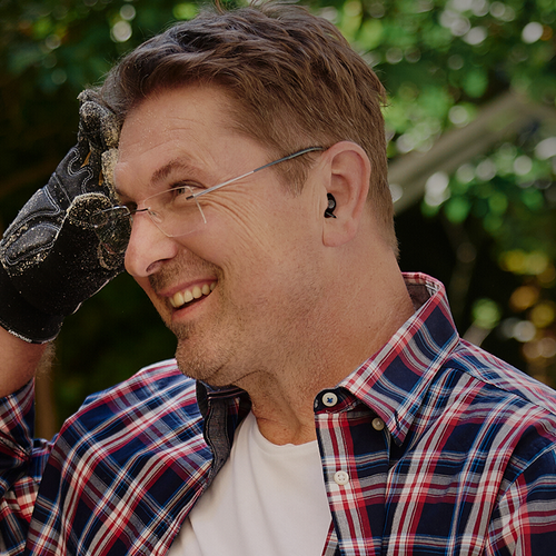 Mann mit schwarzen Gehörschutzstöpseln, Schutzbrille und Handschuhen, arbeitet draußen und lächelt.