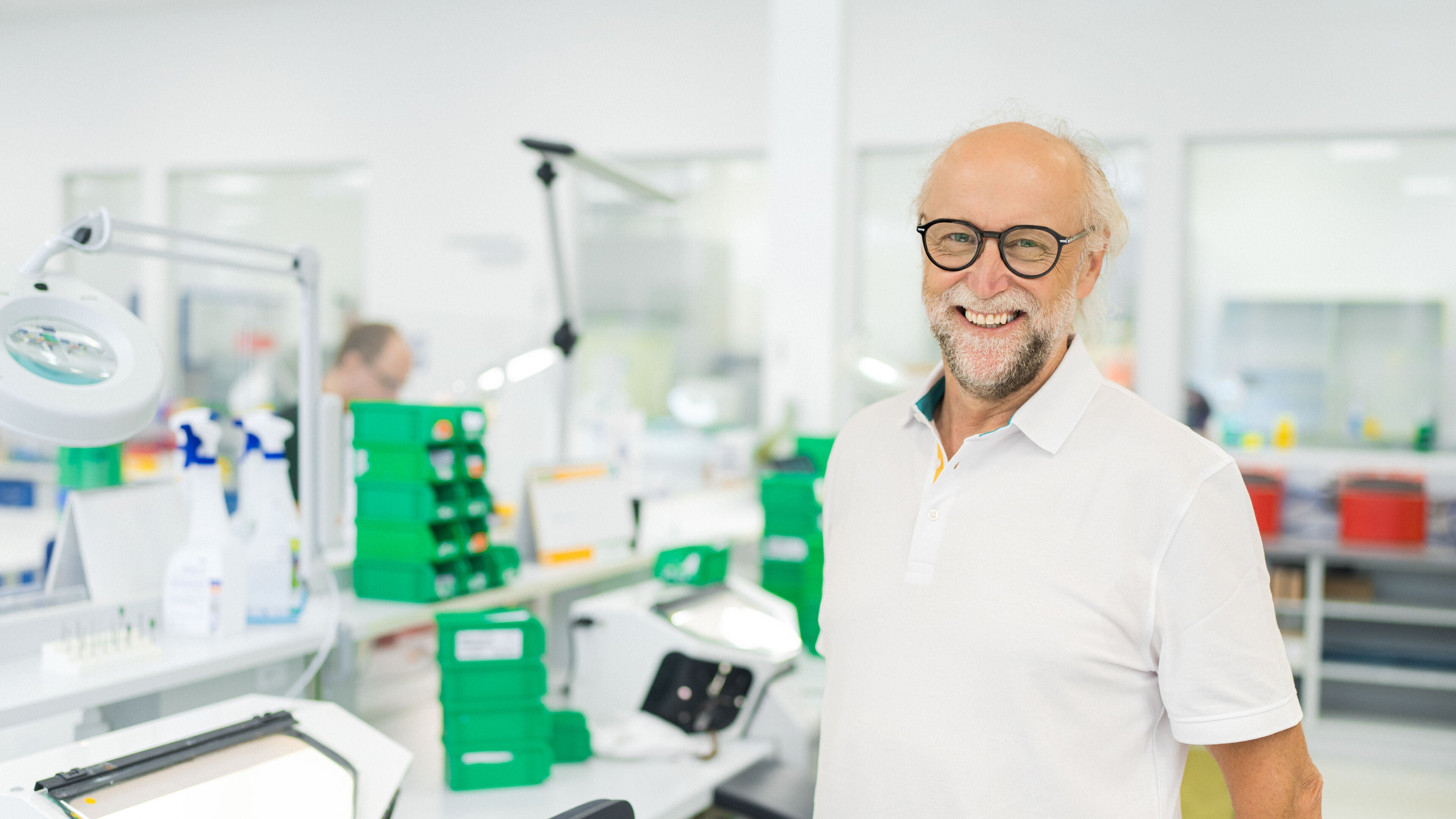 Freundlicher älterer Mann mit Brille und weißem Polohemd lächelt in einer modernen Fertigung von Neuroth mit technischen Arbeitsplätzen und Werkzeugen im Hintergrund