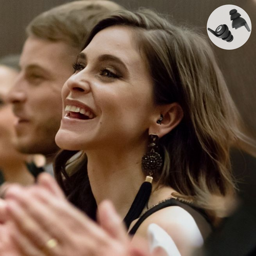 Lächelnde Frau in elegantem Outfit trägt diskrete schwarze Ohrstöpsel während eines Konzerts. Die Ohrstöpsel sind besonders geeignet für Musikgenuss und bieten angenehmen Gehörschutz, ohne die Klangqualität zu beeinträchtigen.