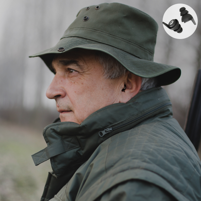 Älterer Mann mit schwarzen Gehörschutzstöpseln, trägt grüne Jagdbekleidung und beobachtet aufmerksam die Umgebung.