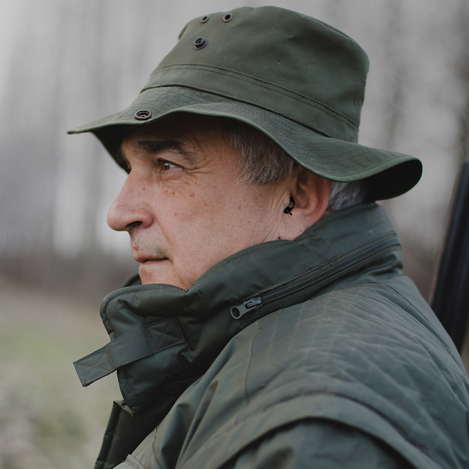 Älterer Mann mit schwarzen Gehörschutzstöpseln, trägt grüne Jagdbekleidung und beobachtet aufmerksam die Umgebung.