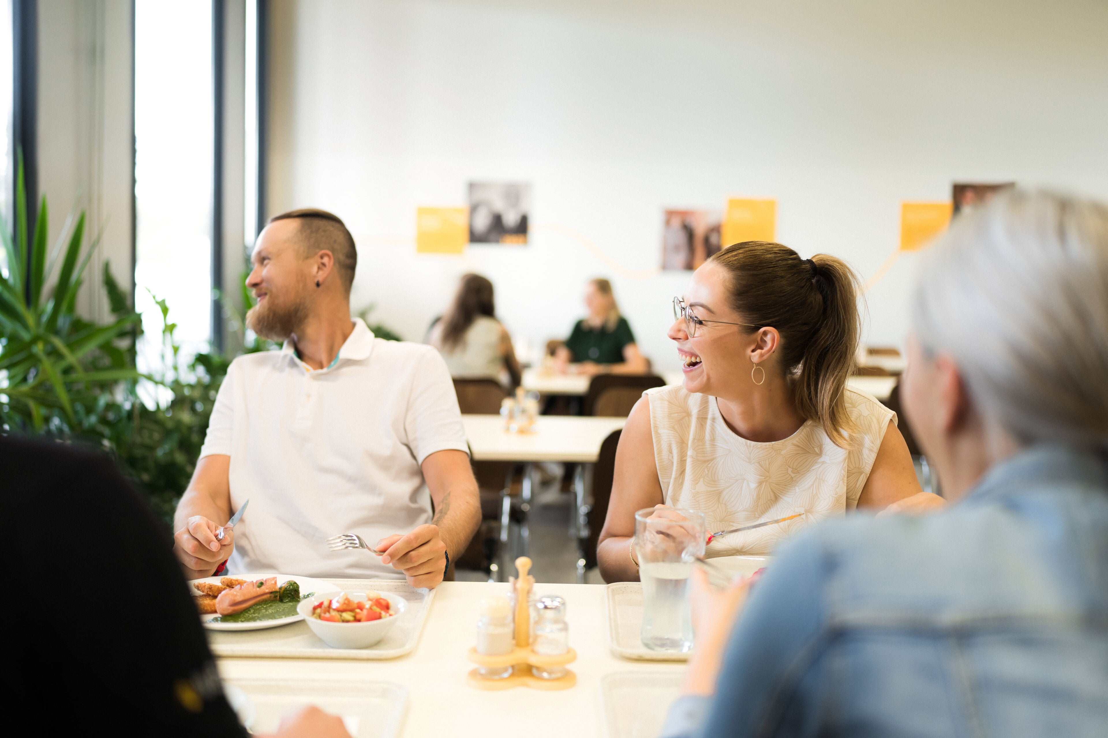 Fröhliche Menschen beim Mittagessen in einer Kantine – Angenehme Gespräche in lebhafter Umgebung, in der hochwertige Silikon-Ohrstöpsel mit Filter für angenehme Geräuschdämmung sorgen können.