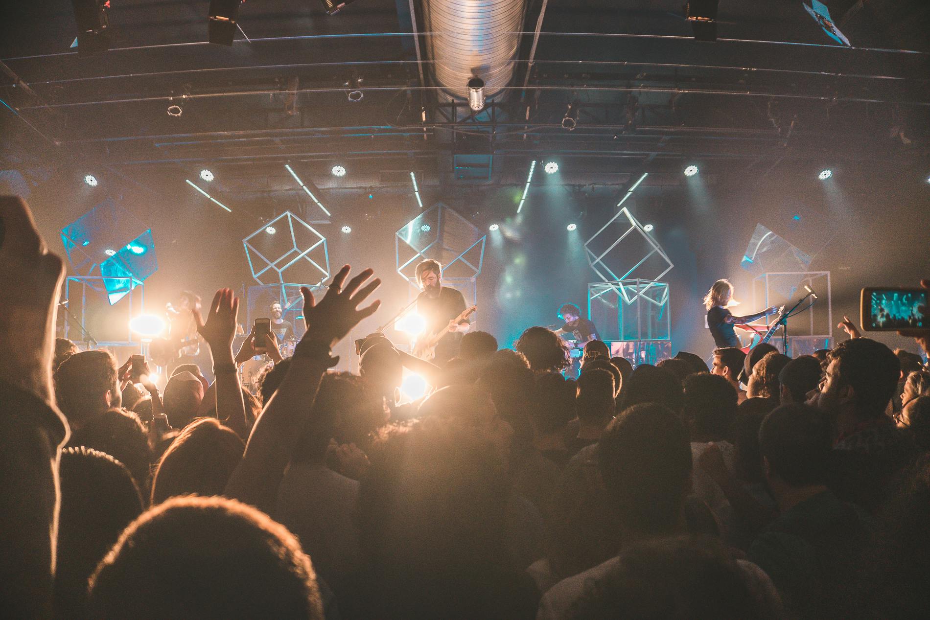 Menschenmenge bei einem Live-Konzert mit Band auf der Bühne, stimmungsvolles Licht und erhobene Hände im Vordergrund.