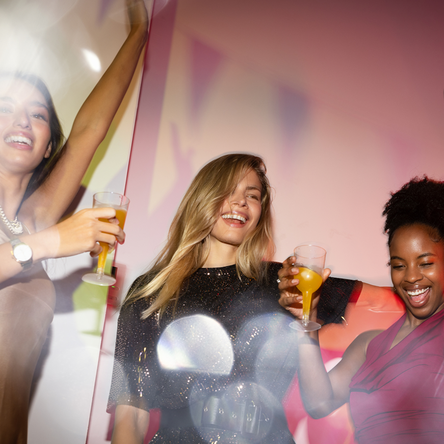  Drei Frauen feiern ausgelassen mit Drinks auf einer Party – idealer Moment für diskreten Gehörschutz beim Feiern.