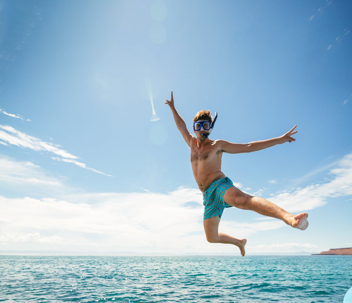 Mann mit Schnorchelmaske springt lachend und dynamisch vom Boot ins blaue Meer bei klarem Himmel.